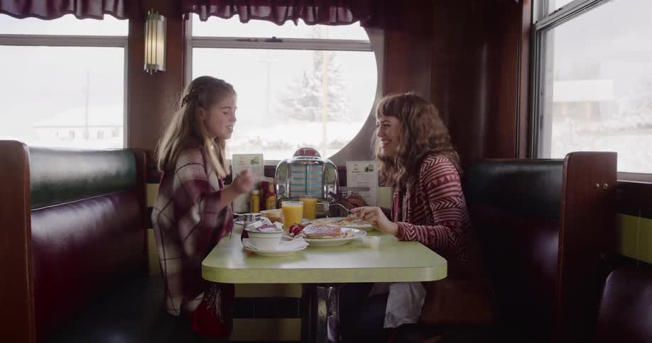 A happy mother and daughter enjoy breakfast together in a cozy, retro diner booth on a snowy winter day