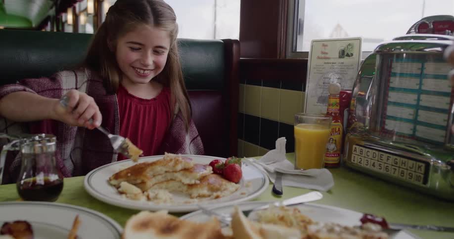 A smiling young girl enthusiastically cuts into her delicious pancake breakfast at a classic retro diner
