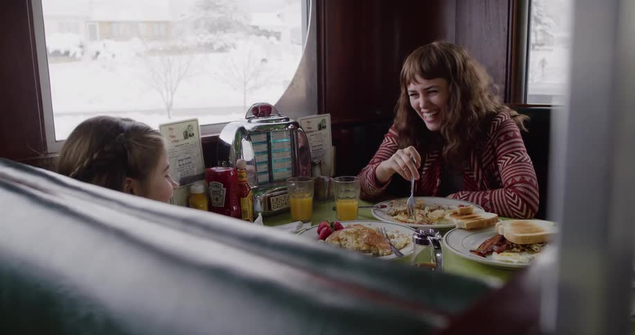 A happy mother and her daughter laugh together while eating breakfast in a cozy diner booth on a snowy winter day