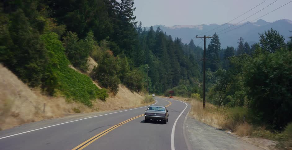 A classic muscle car cruises along a winding mountain road surrounded by a lush green forest