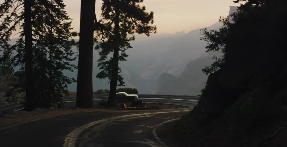 A classic car drives along a winding mountain road revealing the iconic Half Dome of Yosemite National Park at sunset