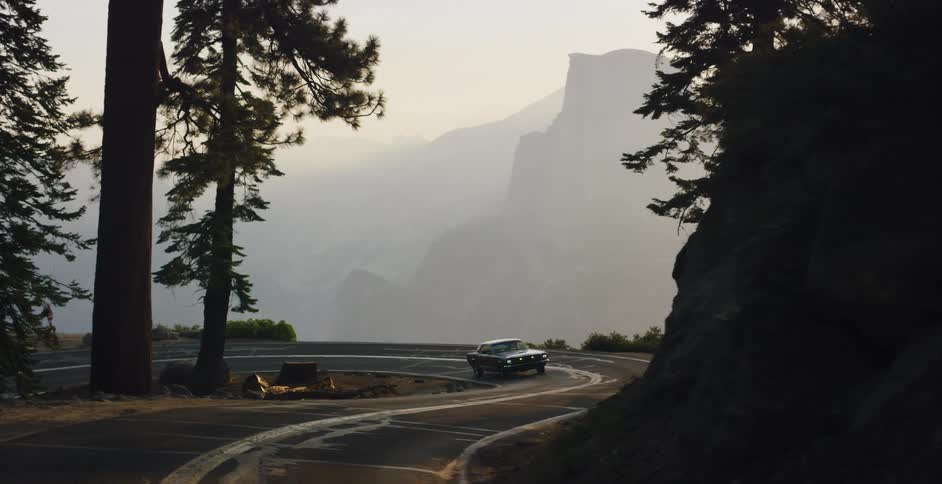 A classic muscle car emerges from the shadows to drive a scenic mountain road overlooking a hazy Half Dome in Yosemite National Park at sunrise