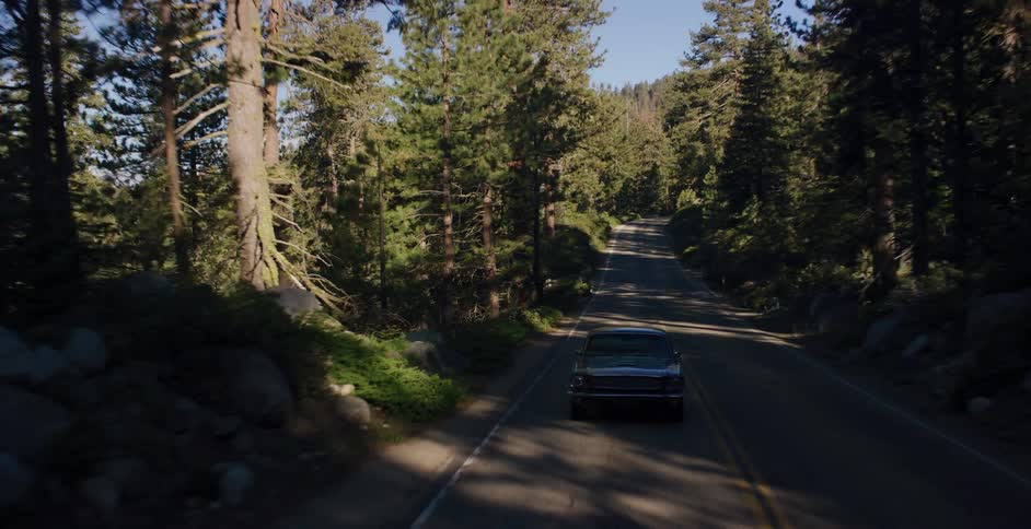An aerial shot pulls back from the trees to reveal a classic black car driving down a winding road through a sunny mountain forest