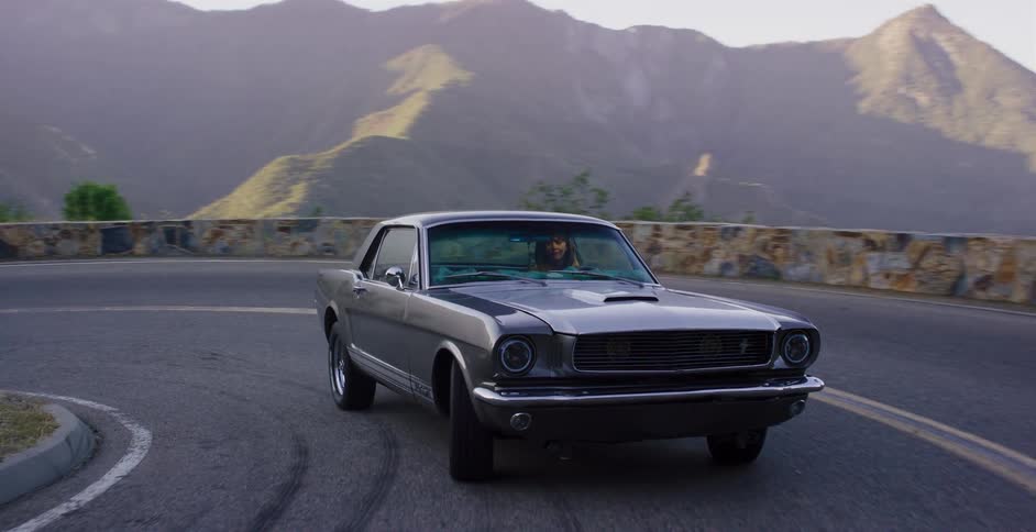 A woman in a classic grey Ford Mustang drives on a winding mountain road in slow motion during a scenic sunset drive