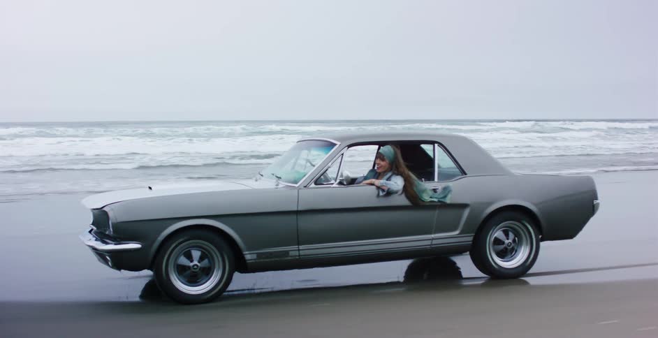 Slow-motion side view of a woman with a headscarf blowing in the wind as she drives a vintage grey Mustang along the shoreline