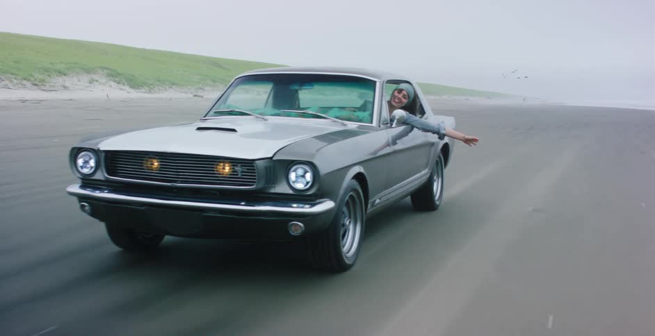 A joyous young woman leans out of a classic Ford Mustang, feeling the wind as it cruises along a vast, sandy beach