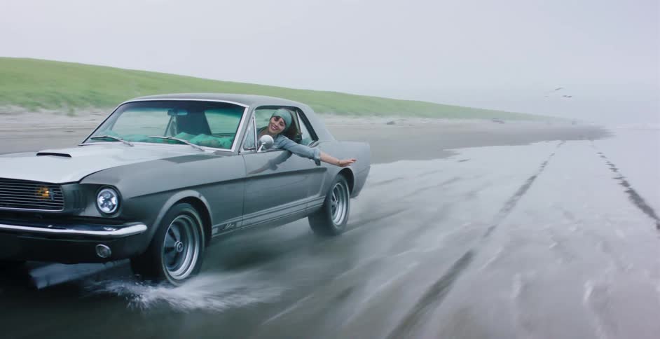 A joyful woman leans out of the passenger window of a classic grey Mustang as it drives along the wet sand of a beach on a foggy day