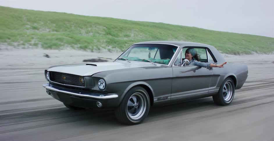Slow motion shot of a carefree woman driving a classic gray Ford Mustang along a sandy beach