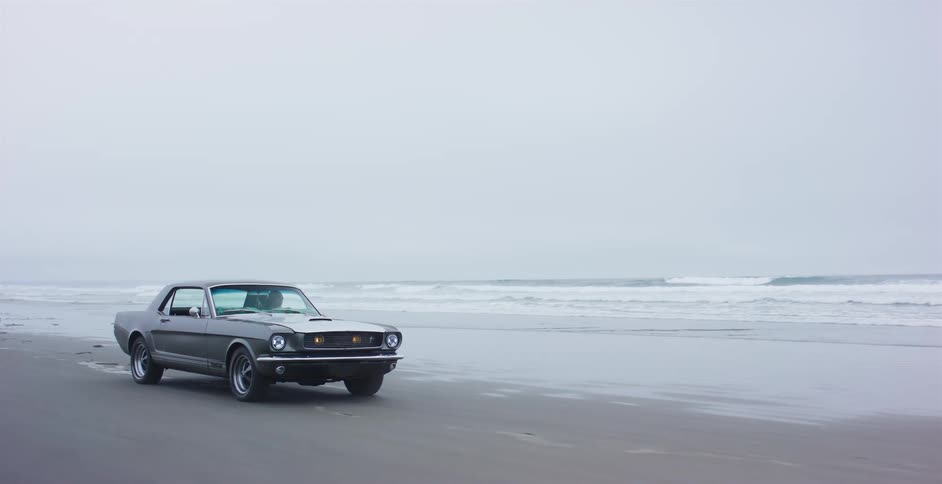 A classic grey Ford Mustang drives along a wet, sandy beach next to the ocean on an overcast day