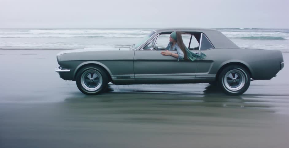 A smiling young woman leans out the window of a classic Ford Mustang as it drives along the reflective, wet sand of an ocean beach