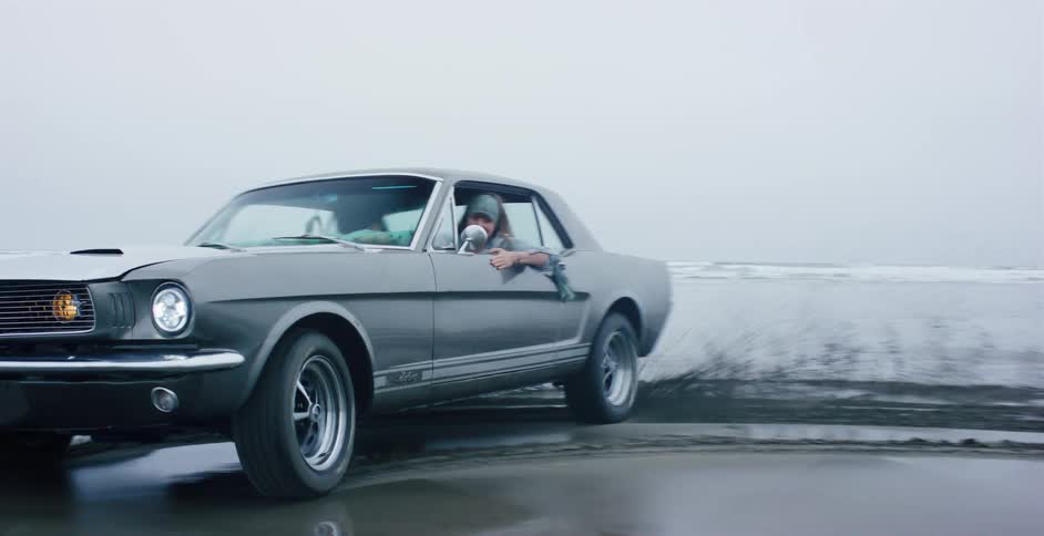 A carefree woman smiles while driving a vintage grey muscle car on a foggy beach
