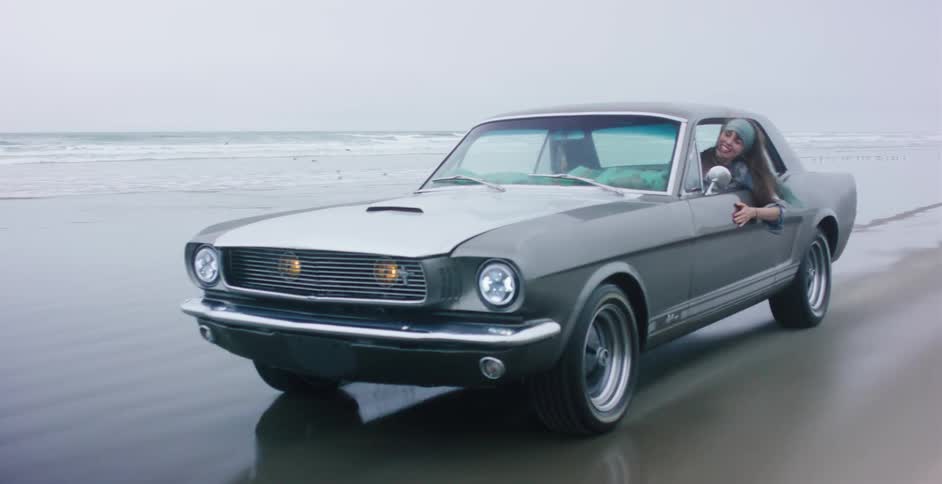 A joyful woman hangs out the window of a classic Ford Mustang driving on the beach in slow motion