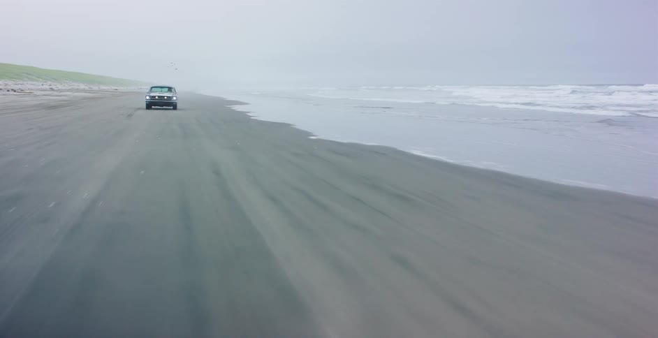 A drone flies backward in front of a lone truck driving down a vast, desolate beach on a foggy, windswept day