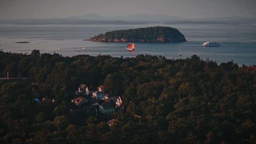 A picturesque view shows a red-sailed schooner and ferry navigating the waters near a forested island and coastal town at sunset