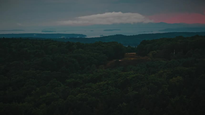 A slow aerial pullback over a dark, dense forest reveals a scenic landscape with a distant ocean and islands under a moody sky at sunset