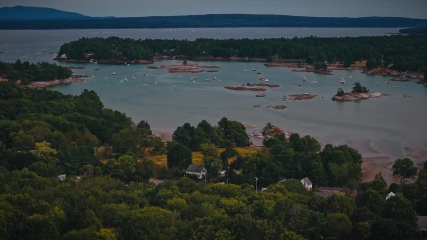 Cinematic aerial pull-back reveals a tranquil bay dotted with sailboats and a quaint coastal town nestled amongst a dense green forest at dusk