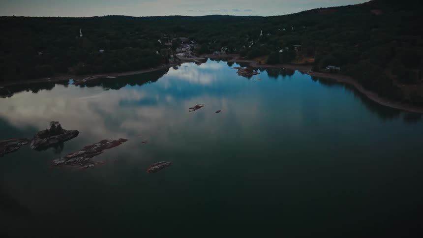 A moody aerial shot slowly pushes over a calm, reflective bay towards a small forested town at dusk