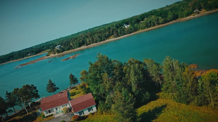 A cinematic Dutch angle aerial shot straightens to reveal a beautiful home on the shore of a serene blue inlet surrounded by lush summer forests