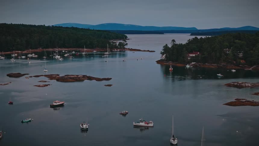 An aerial view slowly pushes in over a tranquil harbor filled with boats at dusk, surrounded by forested islands and distant mountains
