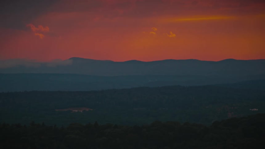 A dramatic red and orange sunset glows over layered mountain silhouettes and a vast, dark forest valley