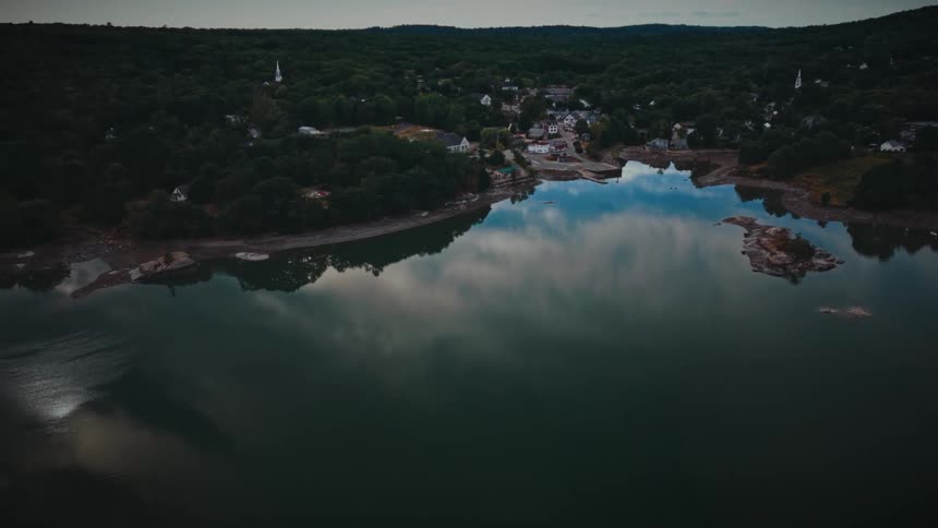 Cinematic aerial footage slowly approaches a small coastal town nestled in a forest, with clouds reflected in the still water