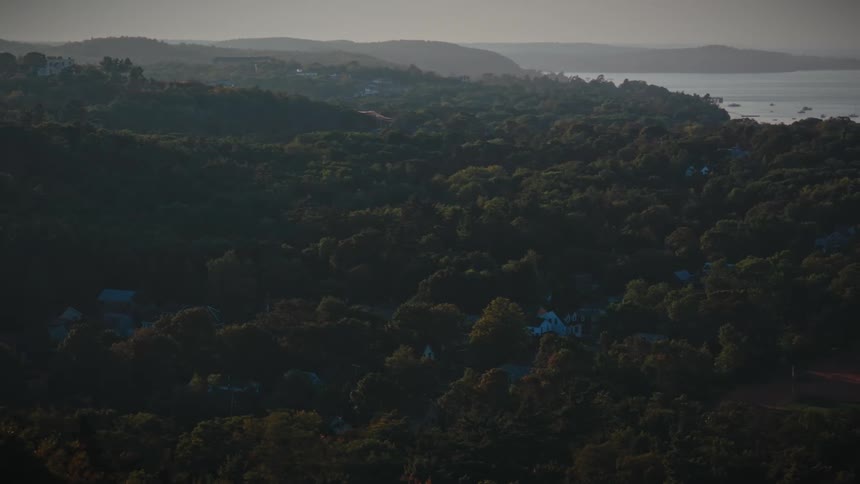 A slow aerial zoom-out reveals a picturesque coastal town nestled within a vast, dense forest at golden hour