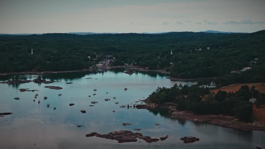 A slow aerial pullback reveals a serene harbor dotted with sailboats, nestled against a densely forested coastline and a quaint coastal town