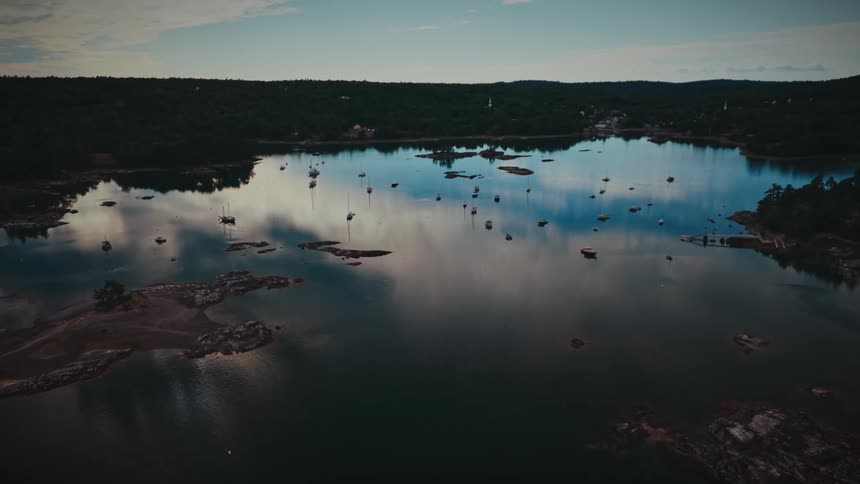 A stunning aerial view captures a quiet harbor with moored sailboats as a lone motorboat slowly cruises across the calm, reflective waters at dusk