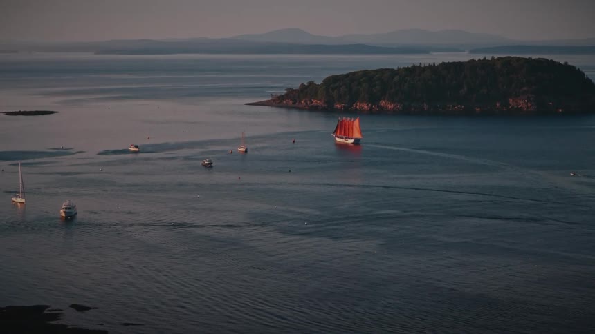 A majestic red-sailed schooner sails through a tranquil coastal harbor surrounded by islands and a forest town during a beautiful sunset