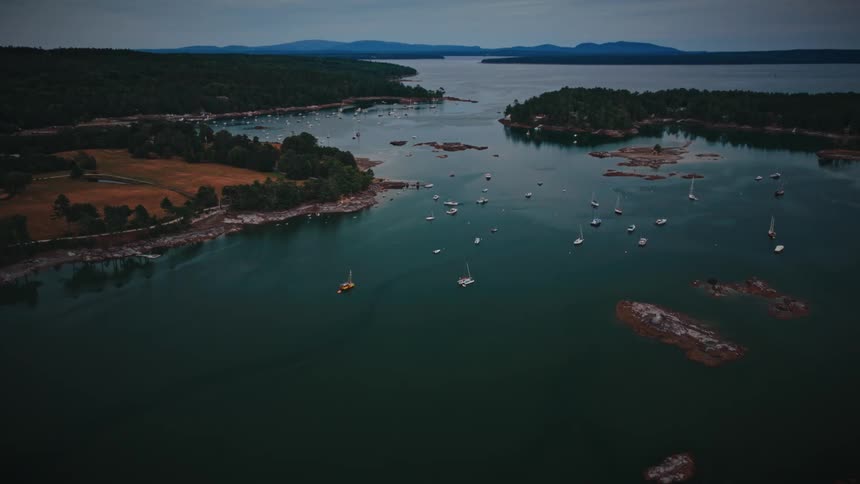 An aerial view pulls back from a tranquil bay dotted with sailboats and surrounded by a dense, rocky, tree-lined coast at dusk