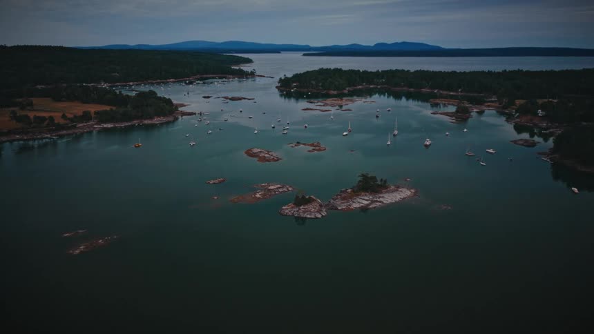 A slow, moody aerial shot flies over a tranquil New England harbor filled with anchored sailboats and surrounded by dense forests at dusk