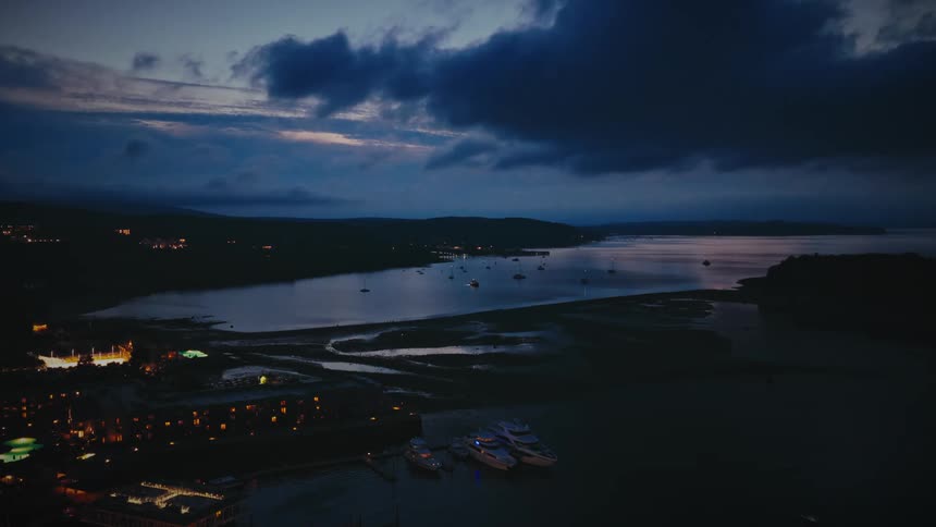 An aerial view of a coastal town and harbor illuminated at dusk as the camera slowly pulls back to reveal the surrounding bay