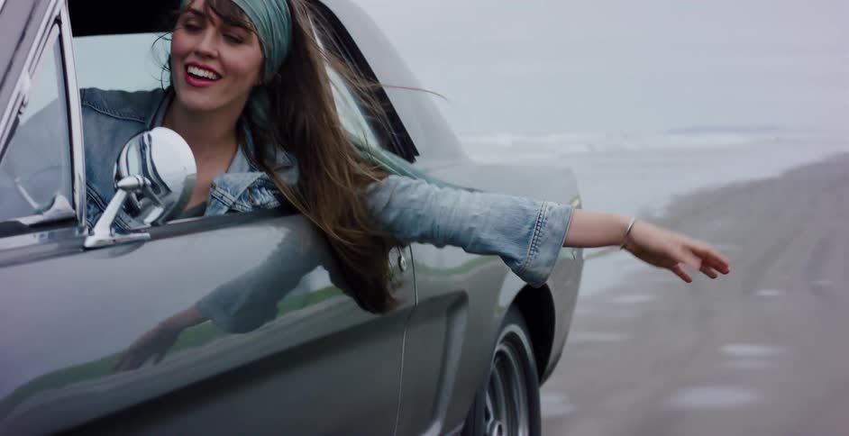A free-spirited woman hangs her arm out the window of a classic car as it drives down a foggy beach