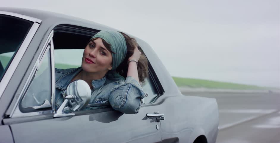 A stylish young woman smiles at the camera while driving a classic grey muscle car along a foggy beach, her hair blowing in the wind