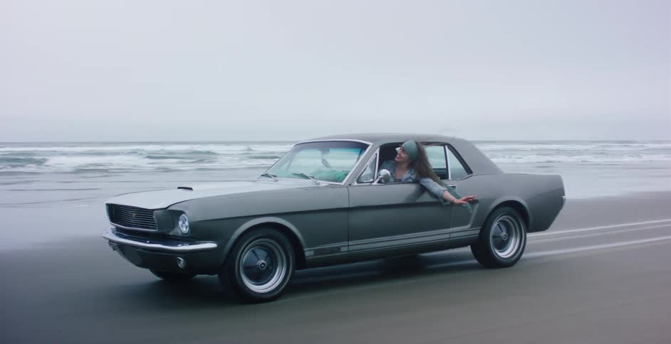 A carefree woman leans out of a classic Ford Mustang, her hair blowing in the wind as they drive along a foggy beach in slow motion