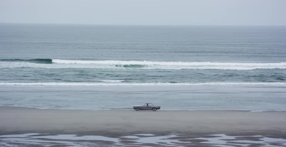 A vintage silver car is parked on a vast, empty beach next to the rolling ocean waves on a cloudy day
