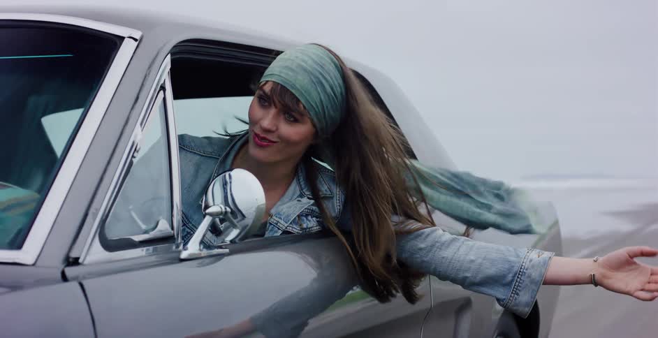 A stylish young woman enjoys a carefree drive along the beach in a classic silver Mustang, with her hand and hair blowing in the wind