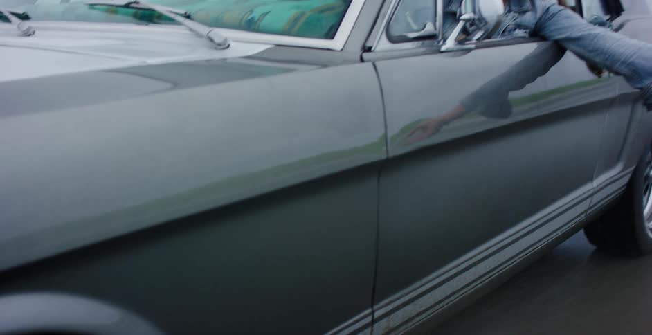 A classic car's tire spins on the wet sand before the camera pans up to a carefree young woman driving along the beach with her arm out the window