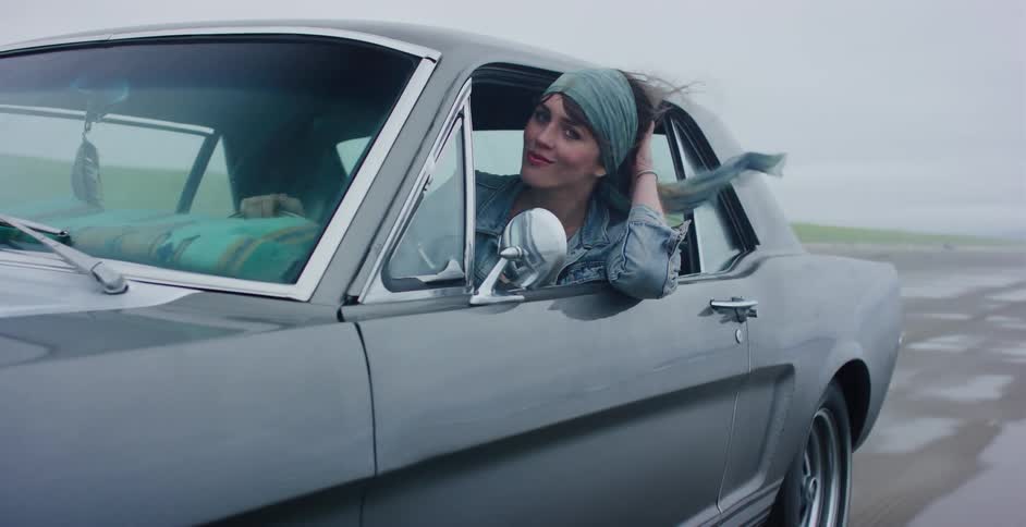 A cool, stylish woman with a bandana in her hair smiles as she drives a classic Ford Mustang along a wet, sandy beach on a windy day