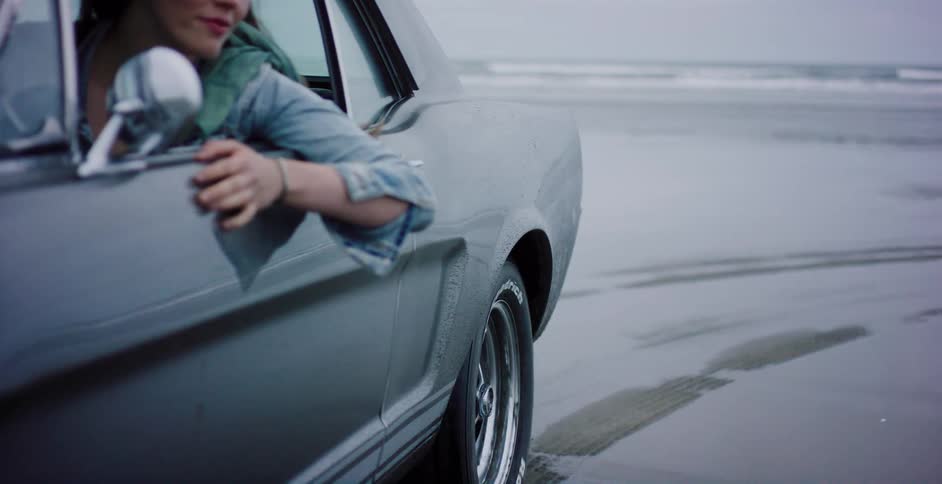 Cinemagraph of a classic car's wheel slowly turning while parked on a wet beach, with a woman's arm resting out the window
