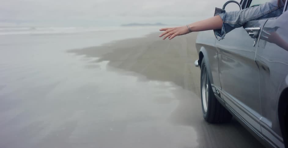 Slow motion shot of a woman's arm catching the breeze from the window of a classic car driving on a beach