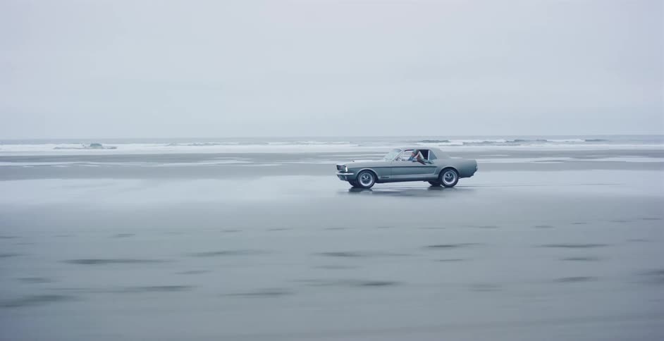 A classic silver Ford Mustang drives along the wet sand of a desolate beach on an overcast day