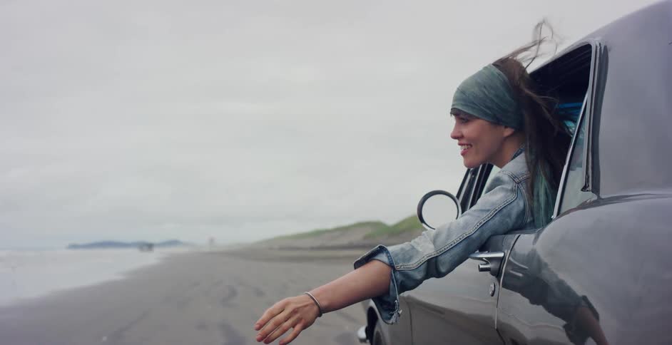 A smiling woman with windblown hair leans out of a car window, enjoying a slow-motion drive on a sandy beach