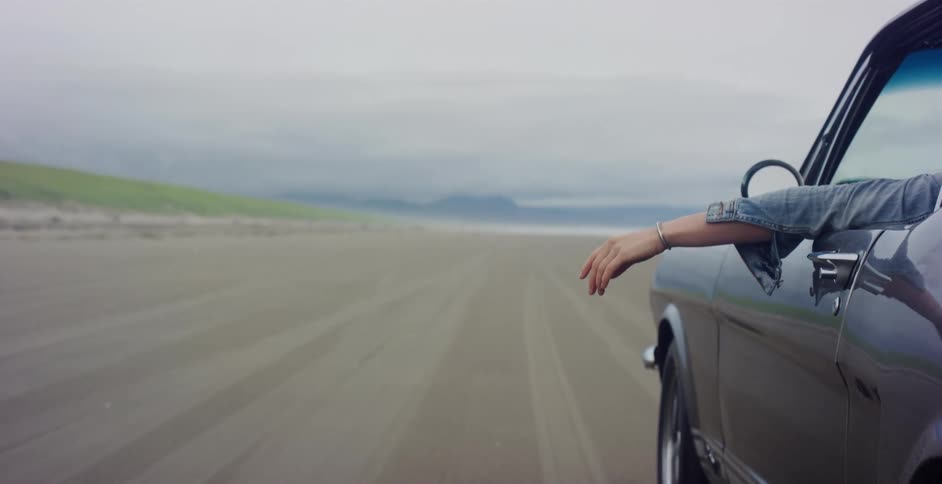 A person's hand catches the wind as they drive a vintage black convertible along a wide sandy beach on a cloudy day