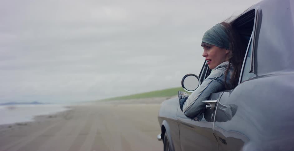 A free-spirited woman with her hair blowing in the wind looks out the window of a classic car as it drives along a sandy beach
