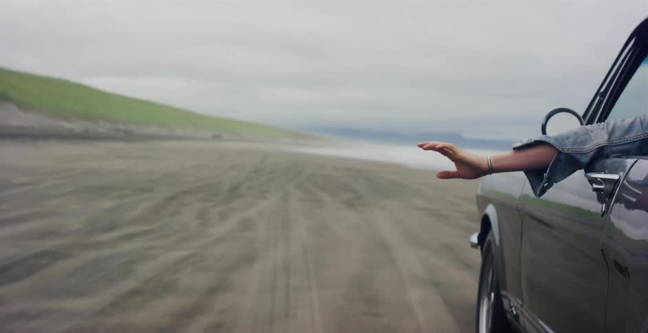 A passenger's hand catches the wind as they drive in a classic black car along a vast, sandy beach on an overcast day