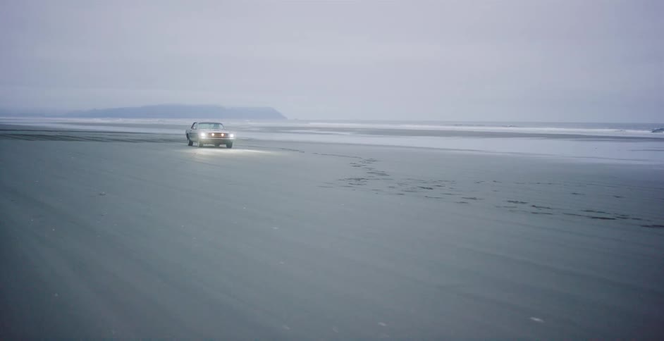 A lone car with headlights on drives across a vast, empty beach on a foggy, overcast day