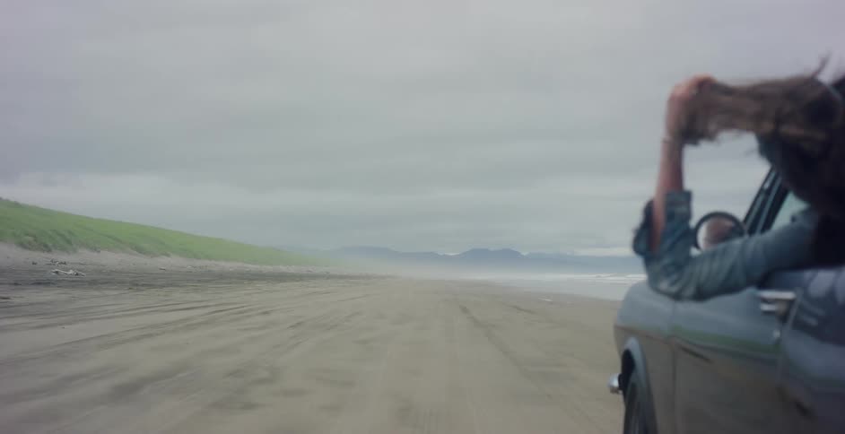 A woman leans out of a vintage car window, her hair blowing in the wind as they drive down an expansive, overcast beach in slow motion