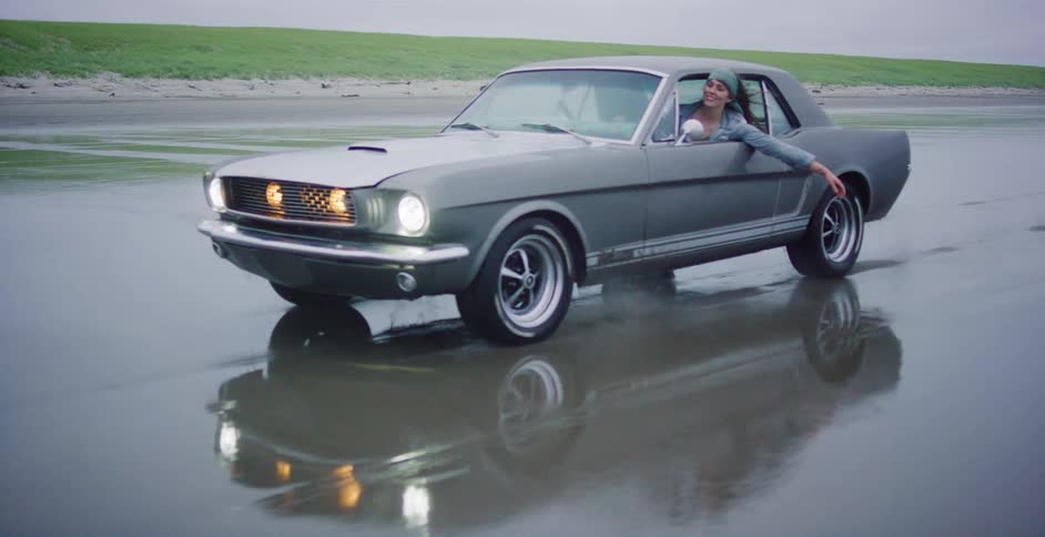 A cool woman in a vintage Ford Mustang enjoys a carefree drive along the wet sand of a beach in slow motion