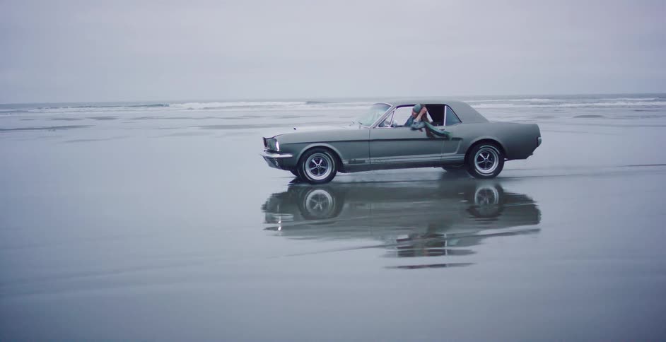 A classic gray Ford Mustang drives along a vast, wet beach, its reflection shimmering on the sand as a passenger relaxes with their feet out the window